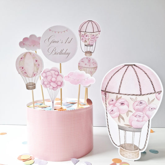 This is a handmade, pink floral hot air balloon cake topper set. It includes balloons, clouds, a personalised topper, and a standing figure. It's made in the UK.