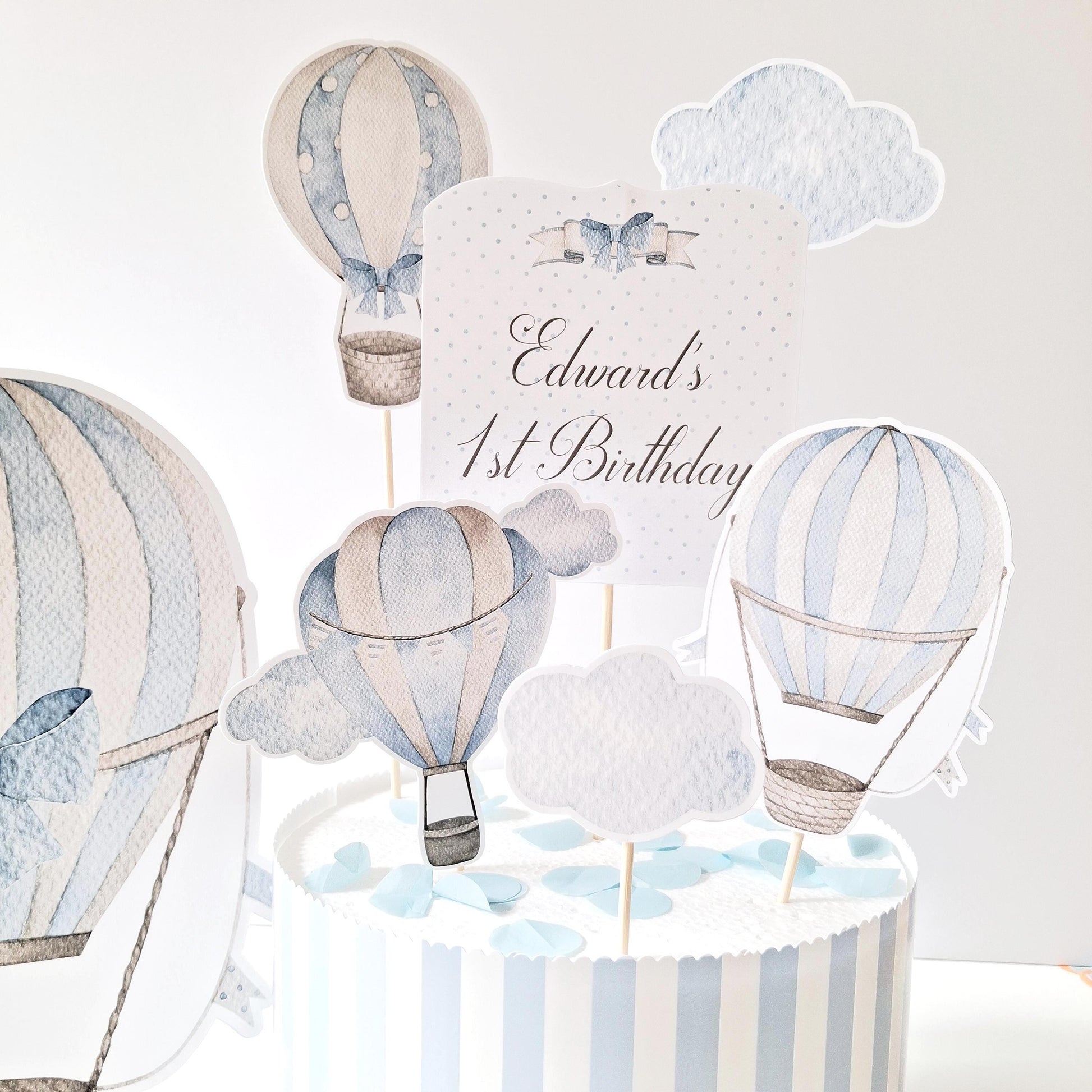 A handmade, soft blue cake topper set from Confites UK. It includes a hot air balloon, clouds, a name-and-age topper, and a matching table figure.