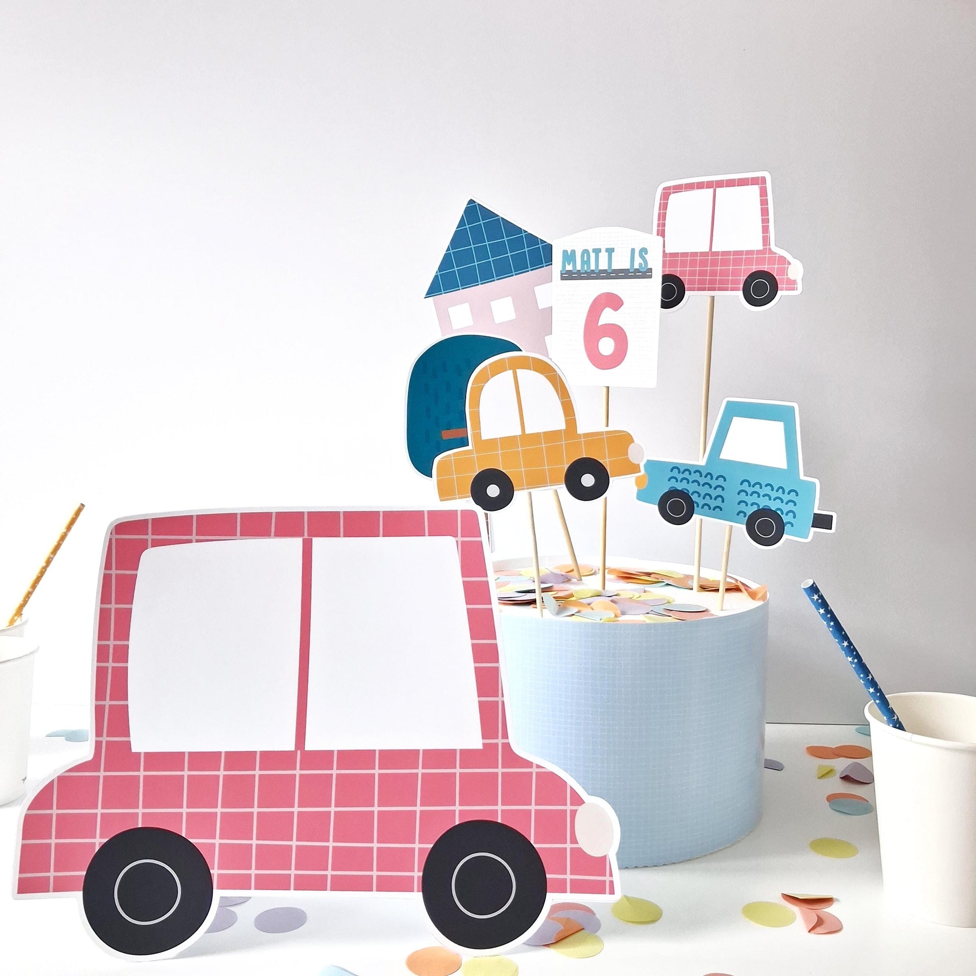 A handmade cake topper set from Confites UK. It includes a custom name-and-age topper, a standing car figure, and additional decorative elements like colorful cars, a house, and a tree.