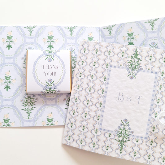 elegant, handmade wedding favours featuring a blue botanical design. Each favour includes chocolate, is tied with a ribbon, and can be personalized with names or dates. They are made in the UK and intended as thank-you gifts for wedding guests.